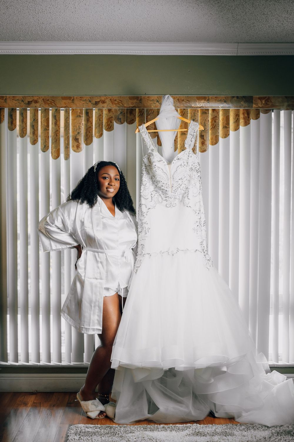 A white mermaid wedding dress hangs elegantly next to someone wearing a bridal robe in a room with vertical blinds.