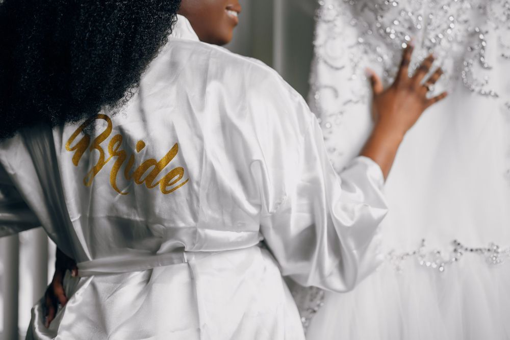 A white satin robe with gold 'Bride' lettering shown from behind as someone reaches toward a wedding dress.