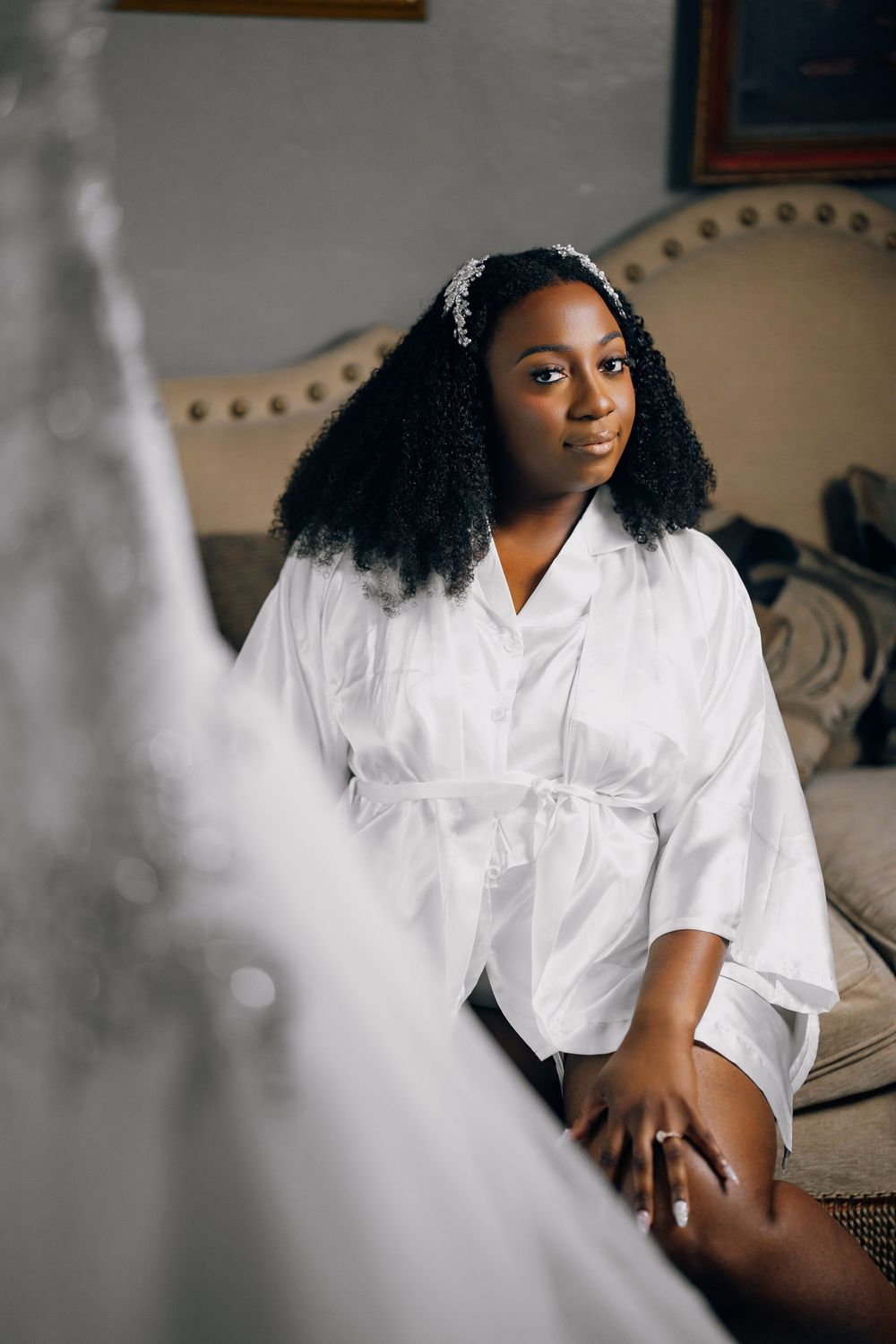 A person in a white silk robe sits on an upholstered chair while getting ready on their wedding day.