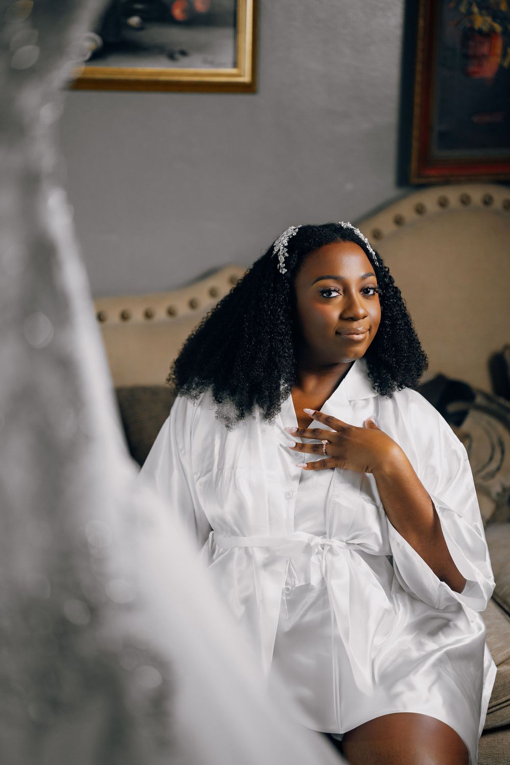 A woman in a white silk robe sitting on an elegant upholstered chair with natural curly hair and soft lighting.