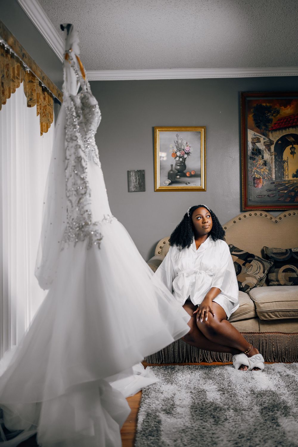 Elegant white wedding gown hanging near a person relaxing in white robe and slippers on couch.