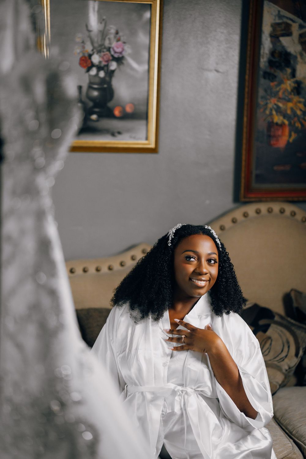 A person in a white robe smiles while getting ready for their wedding day, with framed artwork in the background.
