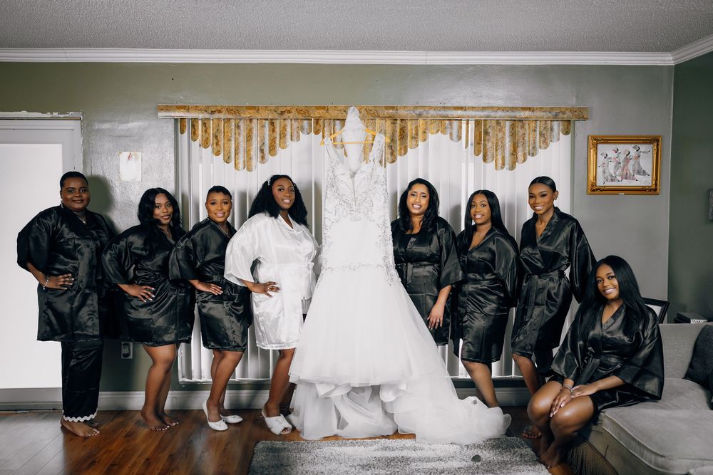 A bridal party in matching black satin robes poses together with a hanging wedding dress in a room.