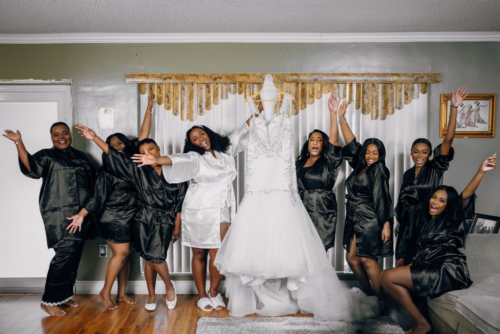 Wedding party members in black robes strike playful poses around a white wedding gown on display.