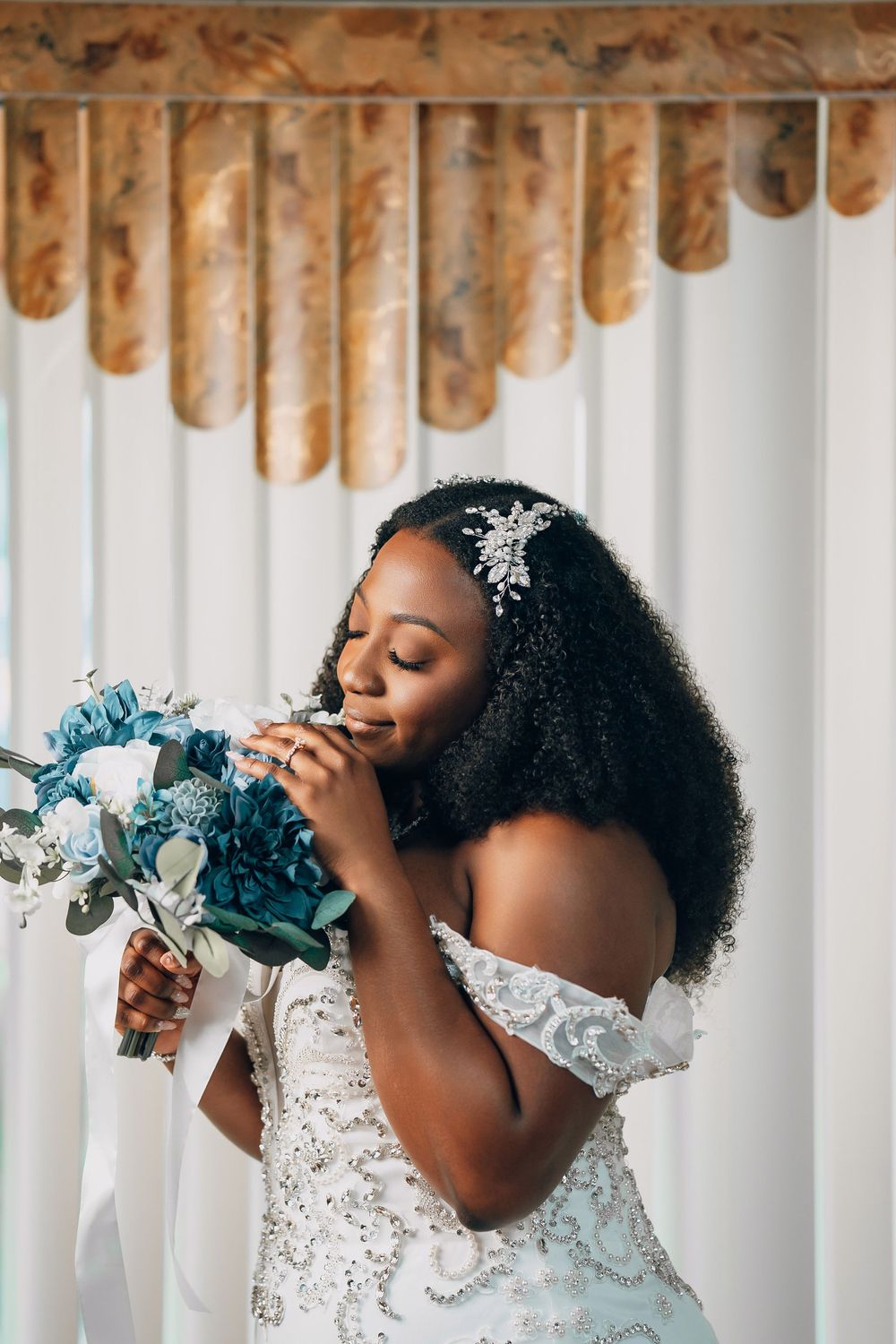 A bride in a white lace gown smells a blue bouquet while standing against a curtained background.
