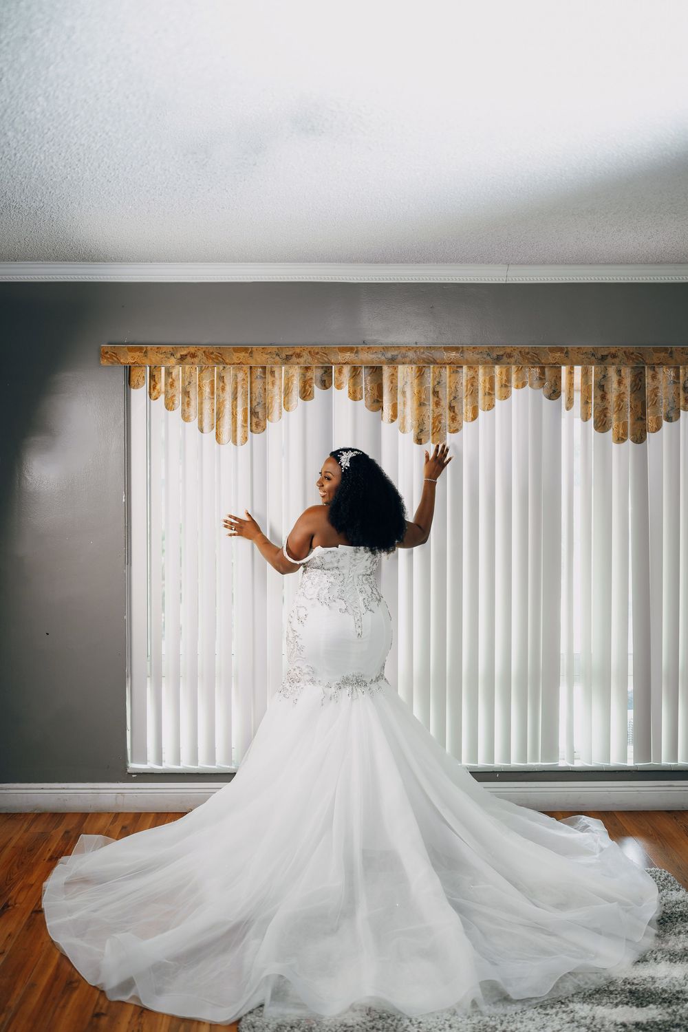 A bride in a stunning white mermaid wedding gown poses dramatically against white vertical blinds.