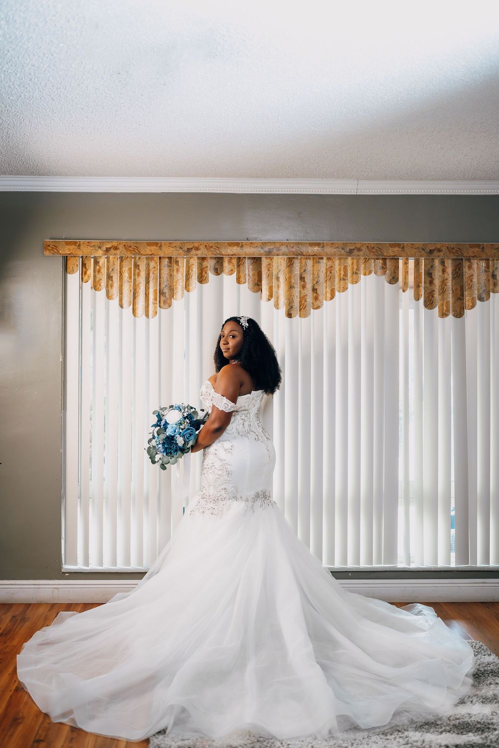 A stunning mermaid wedding gown with a flowing train is showcased against vertical blinds and wooden valance.