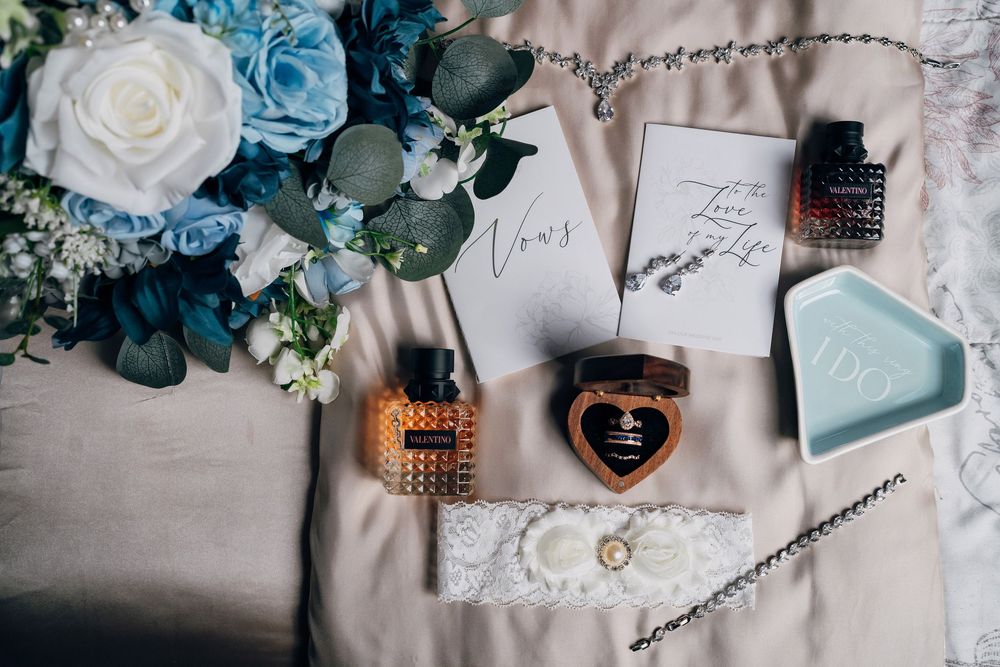 Wedding details flatlay with blue flowers, perfume bottles, and handwritten note on light fabric.