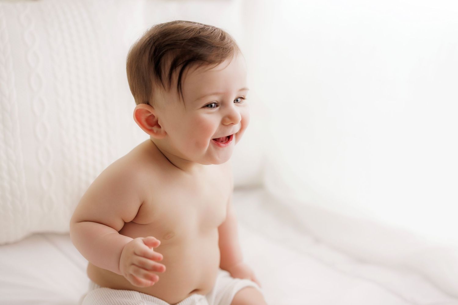 Laughing baby wearing white diaper sitting on light-colored backdrop during professional photo session.