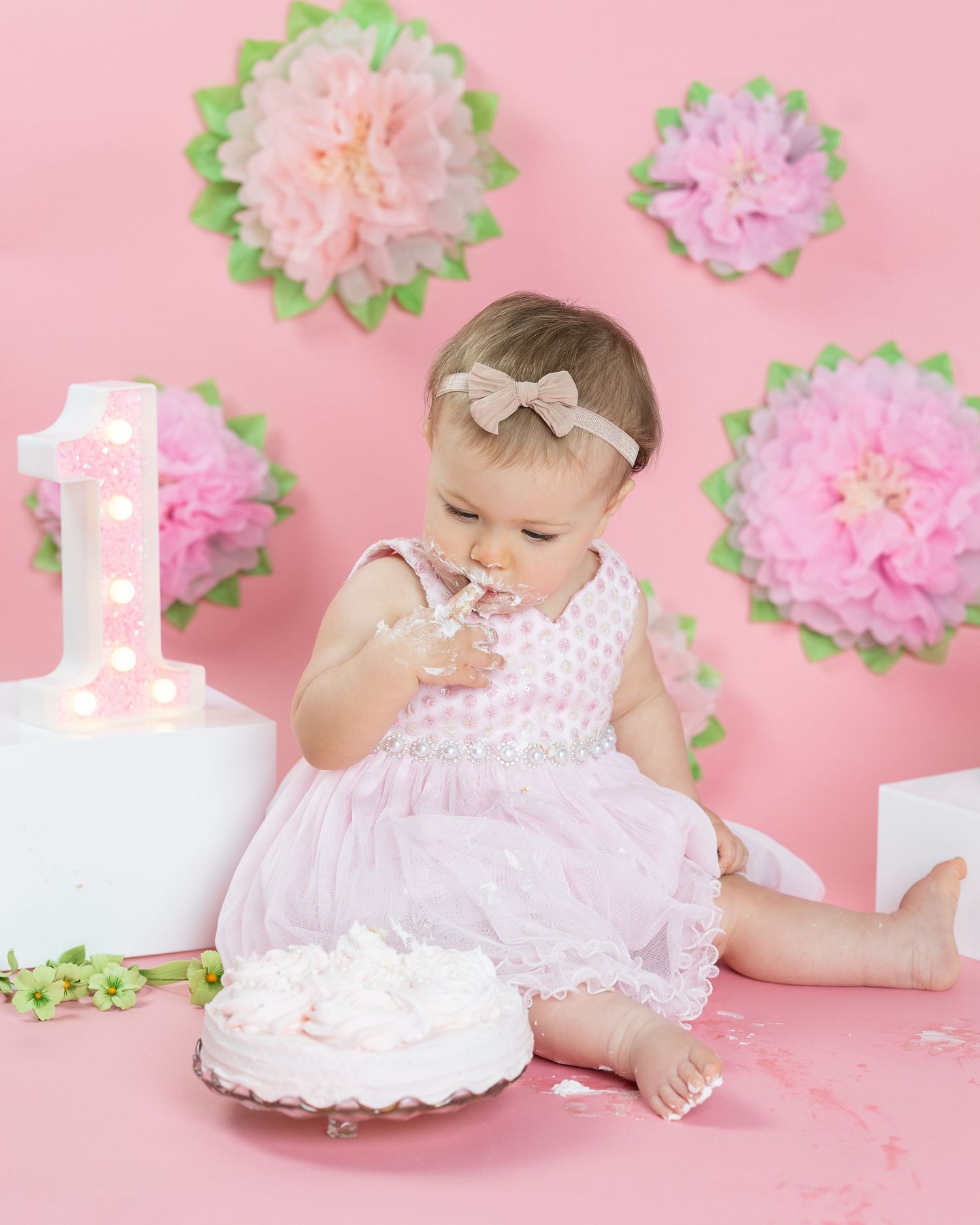 Baby in pink dress celebrates first birthday with cake smash photo session against pink wall with paper flower decorations.