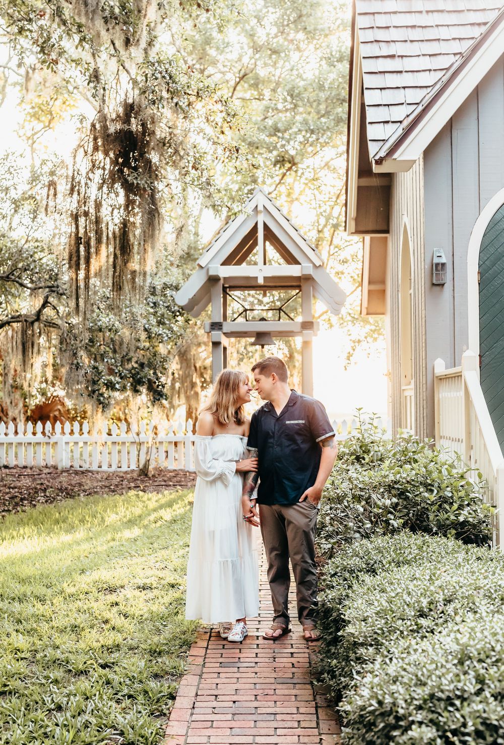 Couple in light-colored outfits standing on a brick path near a charming chapel.