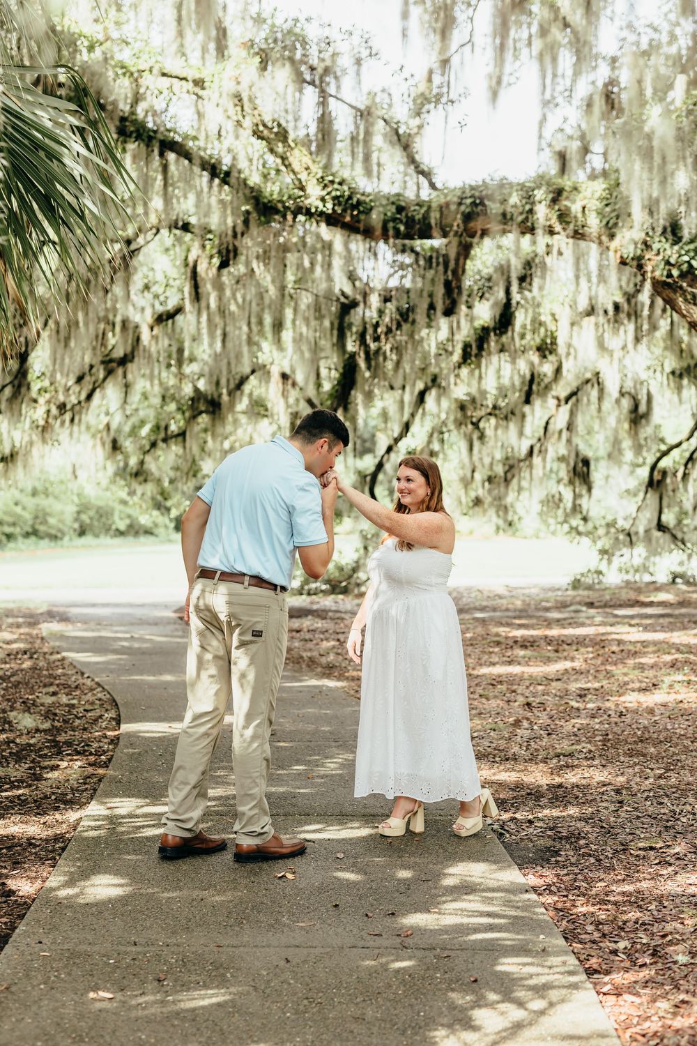 A man kisses a woman's hand on a garden path beneath moss-draped oak trees, in a romantic outdoor setting.
