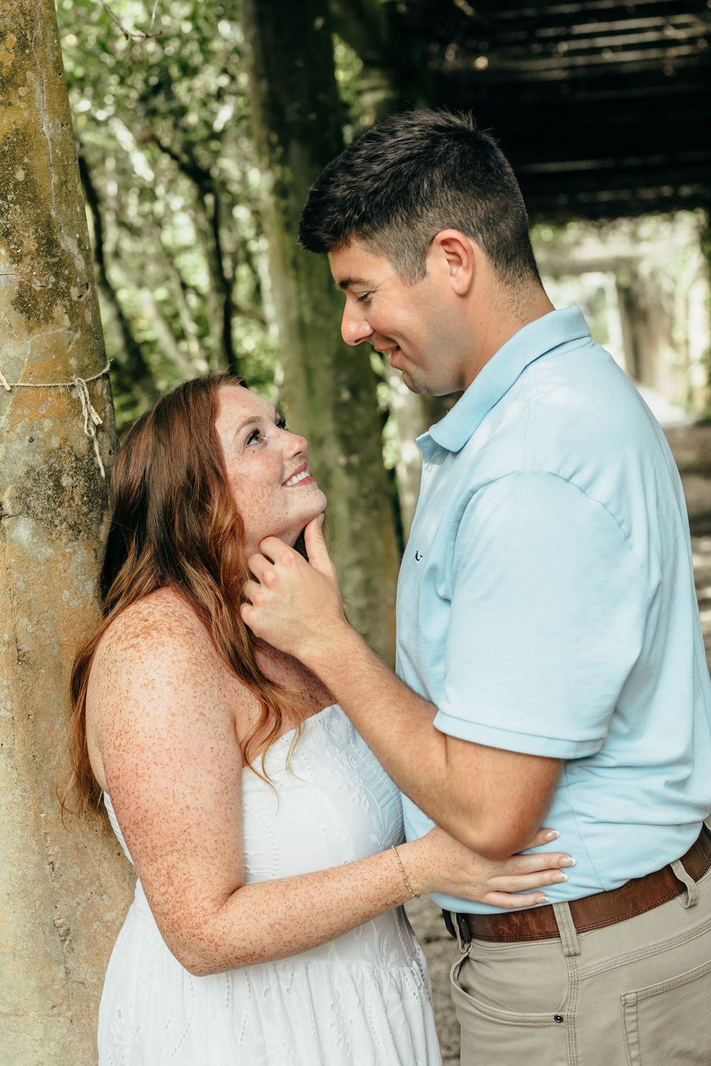 A couple shares a tender moment outdoors, the man gently holding the woman's chin as she gazes up at him lovingly.