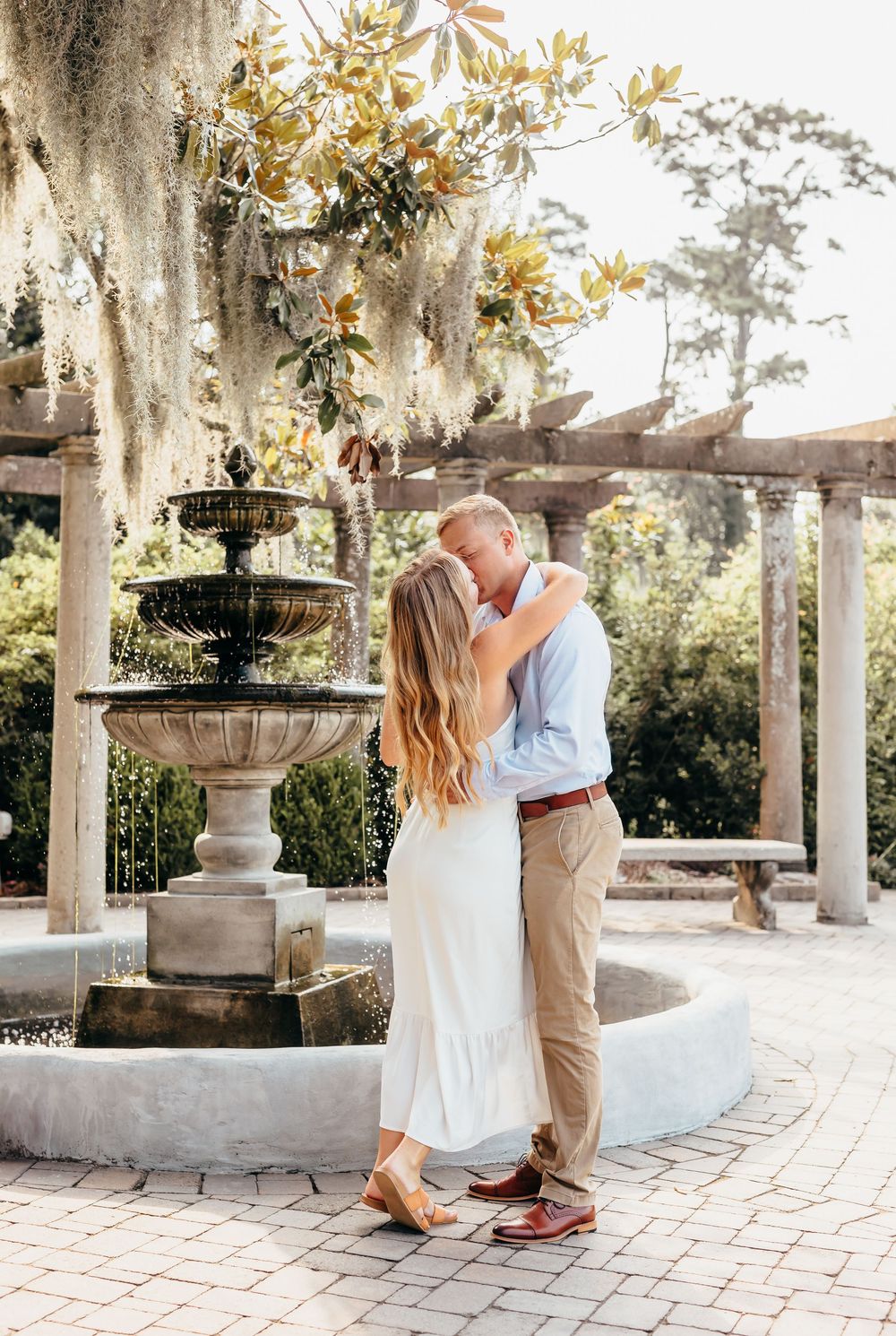 A romantic couple sharing a kiss by an elegant stone fountain with Spanish moss and flowering trees in the background.