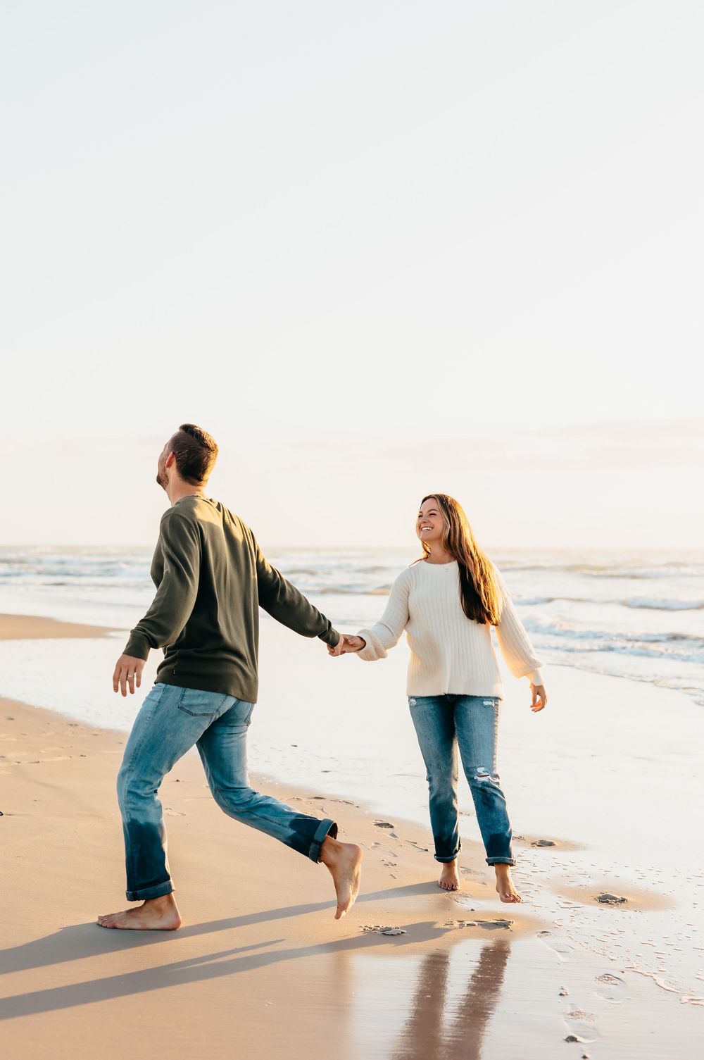 A couple holds hands while dancing playfully on a beach at sunset.