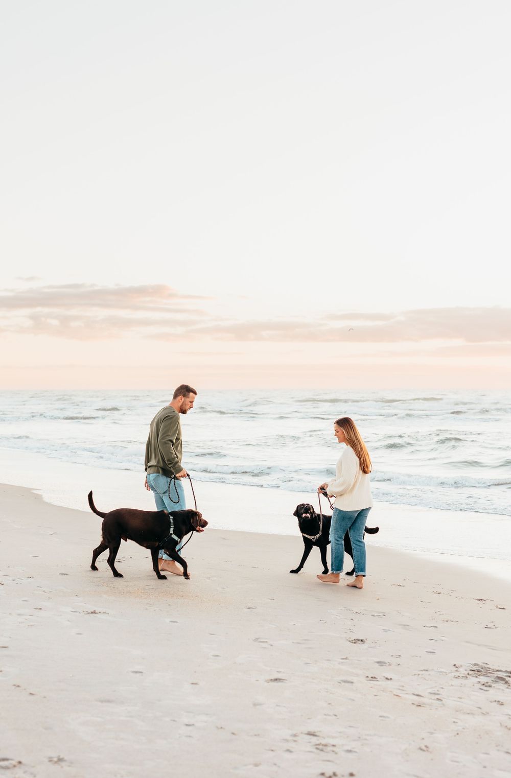 Two people walking their black dogs along a serene beach at sunset.