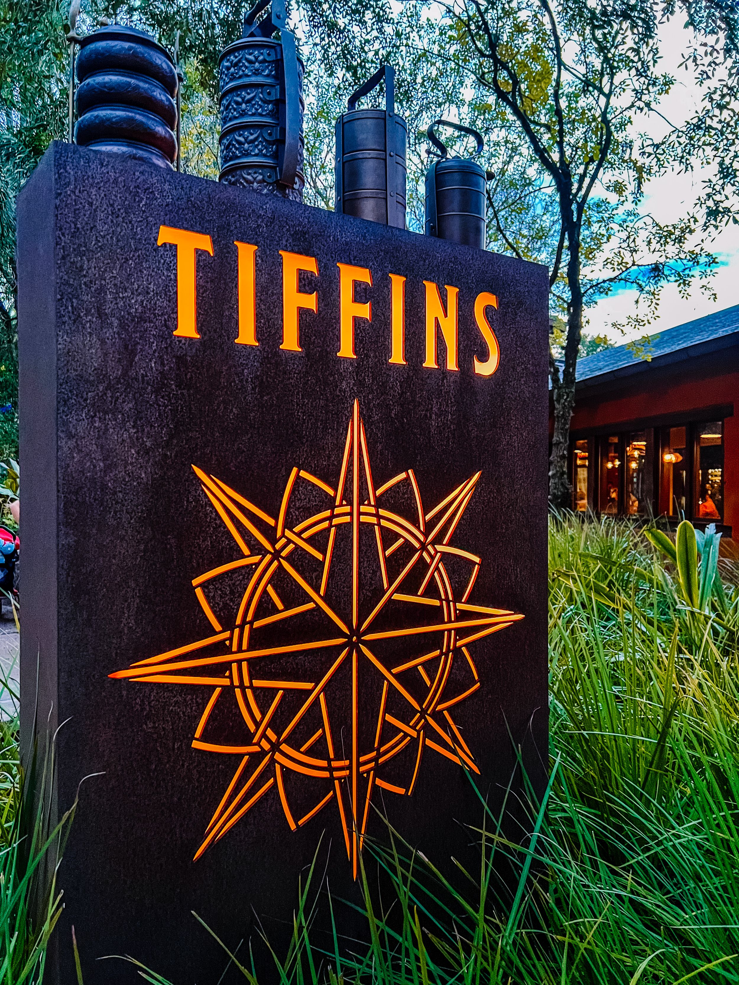 Illuminated Tiffins restaurant signage with geometric star design glowing orange against lush landscaping.