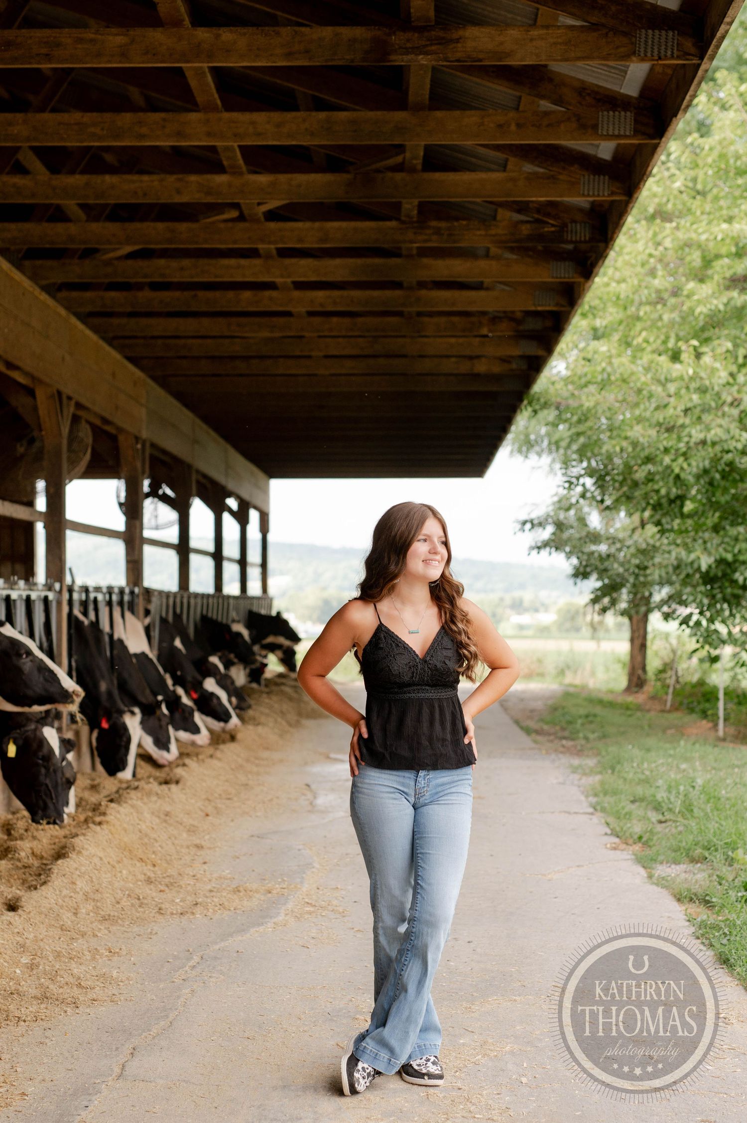 Casual standing pose on path beside dairy barn with rolling hills in background.