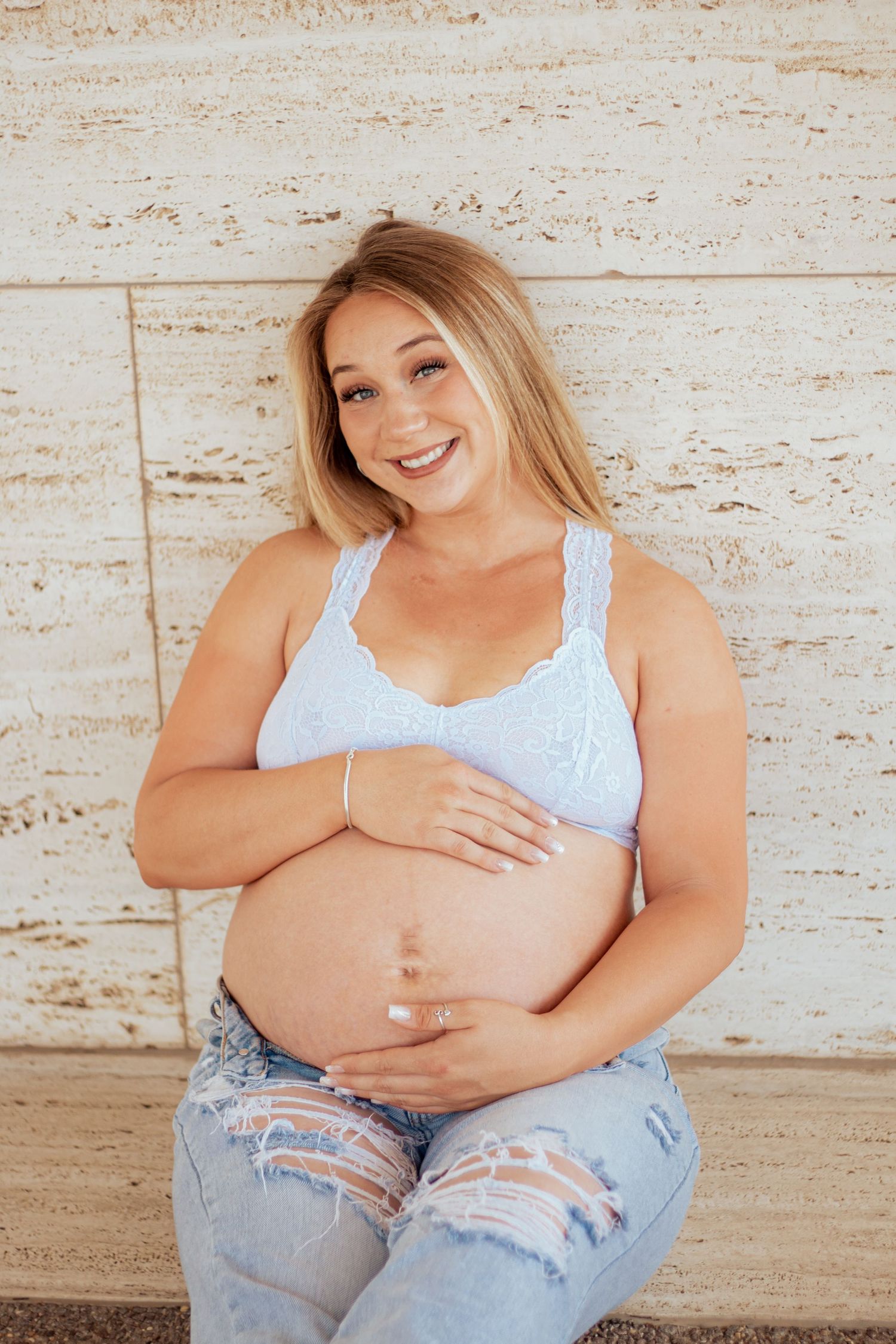 A pregnant woman in distressed jeans and white top poses against a beige wall while cradling her baby bump.