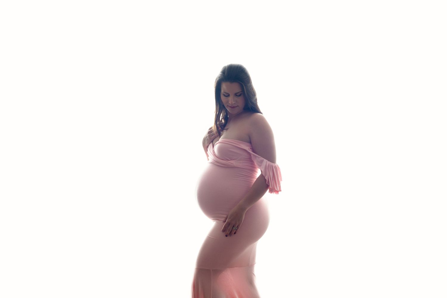 Silhouetted figure in pink dress poses for dramatic maternity photo against light background. Fresno Maternity Photographer