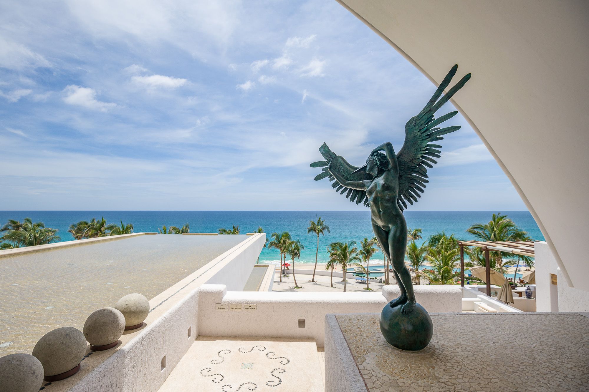Oceanfront terrace with metal human bird sculpture and white architectural arch overlooking beach views. Marquis Los Cabos.