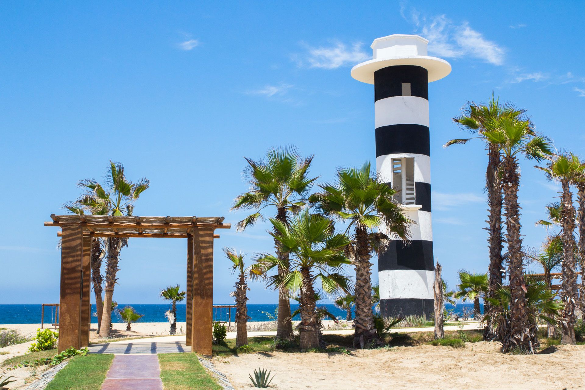 Black and white striped lighthouse tower with palm trees and wooden pergola leading to beach pathway. El Faro by Guaycura.