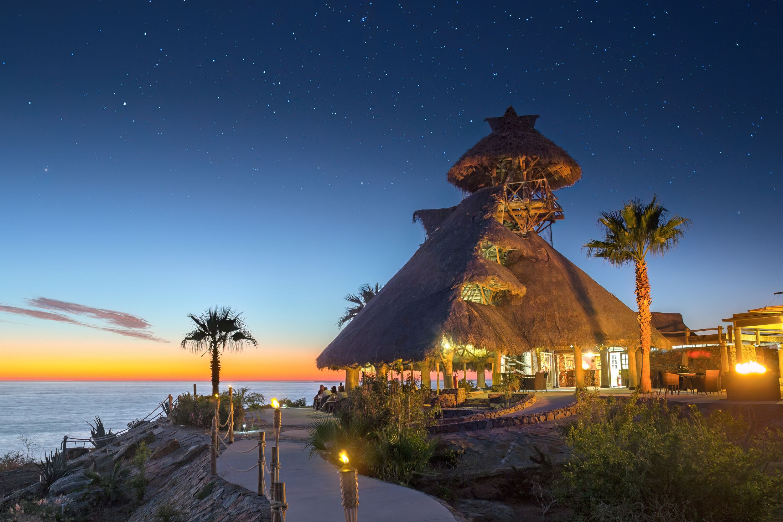 Thatched-roof resort restaurant overlooking ocean at sunset with palm trees and glowing lights. El Mirador by Guaycura.