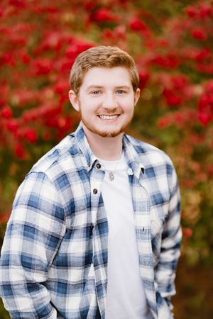 A smiling person in a blue and white plaid flannel shirt stands in front of vibrant red autumn foliage.