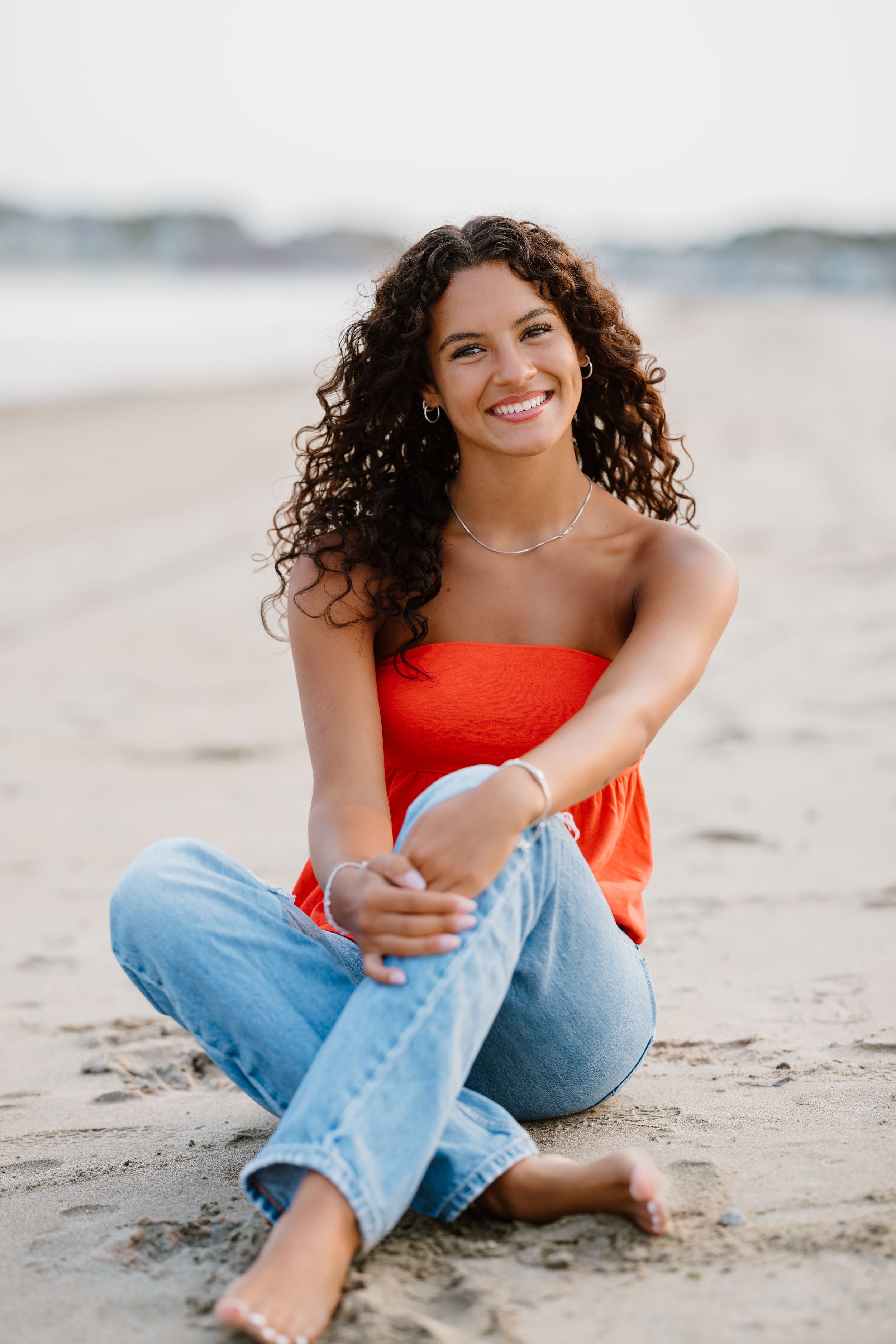 Young woman with curly hair smiling on beach, wearing red tube top and jeans, sitting barefoot on sand.
