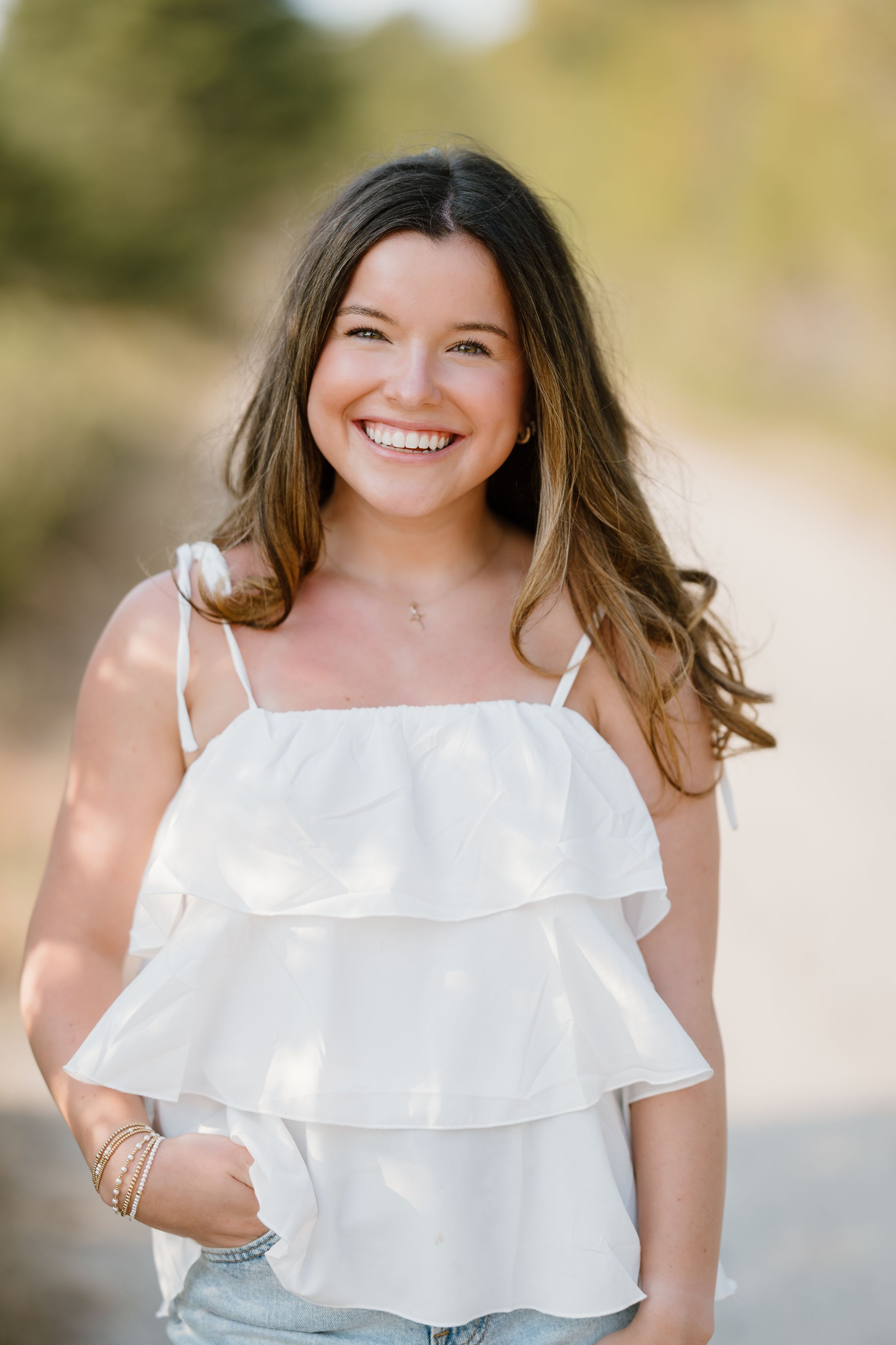 Smiling young woman in white ruffled tank top standing on a sunny dirt path with bokeh background.