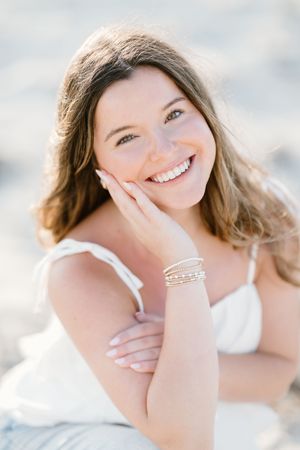 Smiling young woman with brown hair wearing a white dress and bracelets, posing outdoors in natural light.