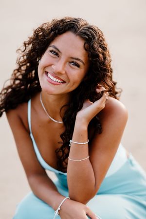 Smiling young woman with curly dark hair wearing a light blue dress and silver jewelry, posing outdoors.