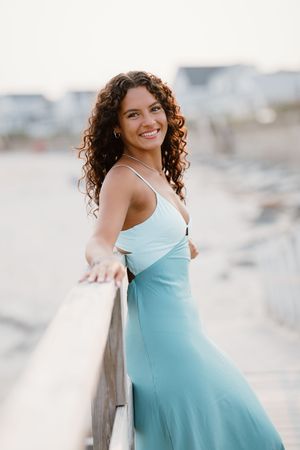 Young woman in a light blue sundress smiling on a beach boardwalk with curly hair and coastal background.