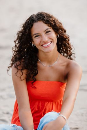 Young woman with curly hair smiling on beach, wearing red strapless top and jeans, silver necklace and hoop earrings.