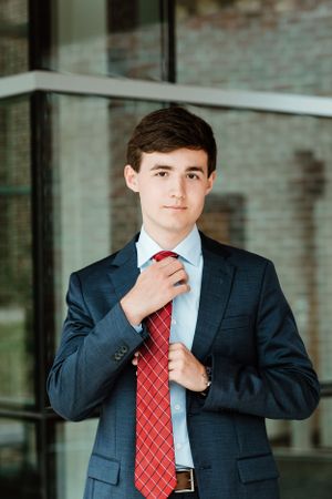 A professional wearing a navy suit and red diagonal striped tie adjusts their collar in a business setting.
