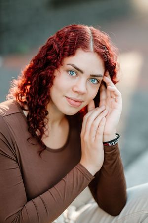 Close-up portrait featuring curly red hair and brown top against a blurred background.