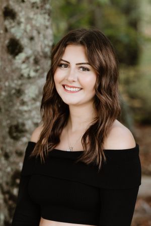 Portrait outdoors against a tree wearing a black off-shoulder top.