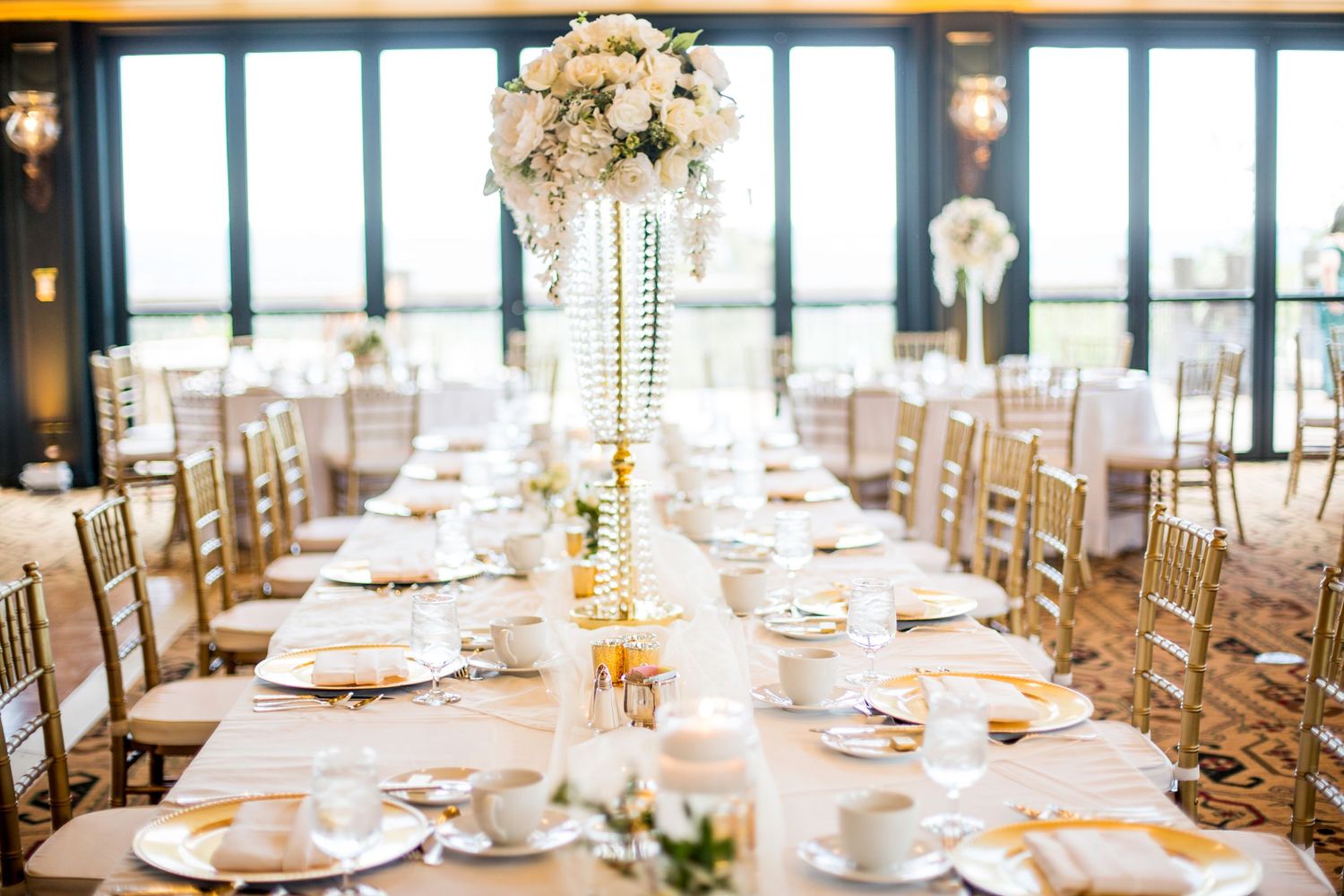 Elegant wedding reception table setting with tall crystal centerpiece and gold chiavari chairs in a sunlit venue.