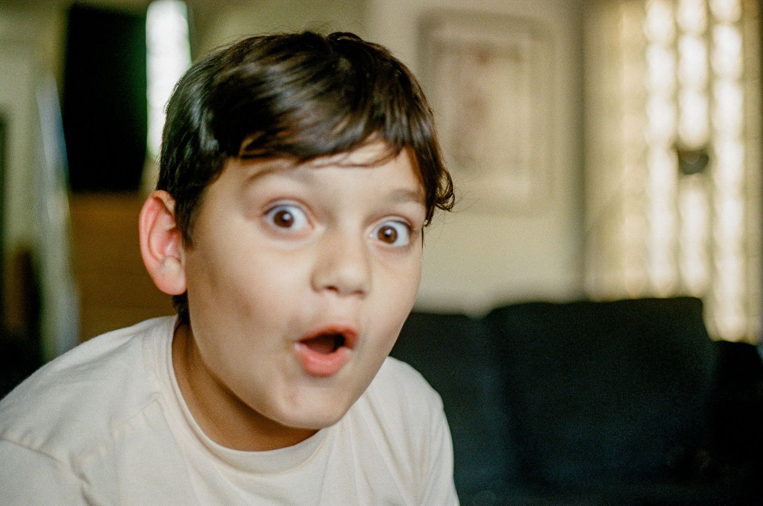 A child with an astonished expression displays wide-eyed surprise while sitting on a dark couch.