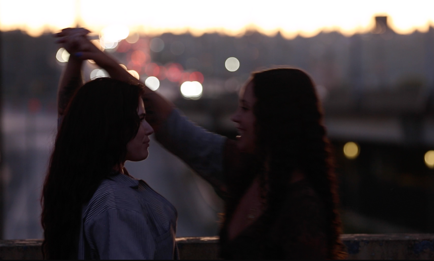 Two silhouettes standing close together at dusk with bokeh lights in the background.