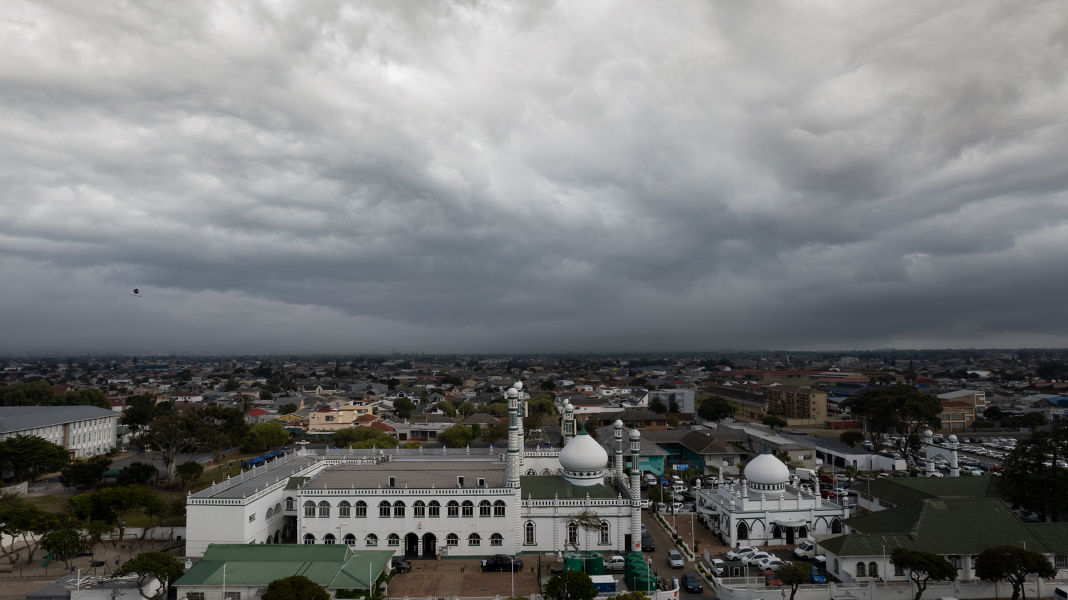 Aerial view of a mosque with white minarets under dramatic stormy clouds in an urban landscape.