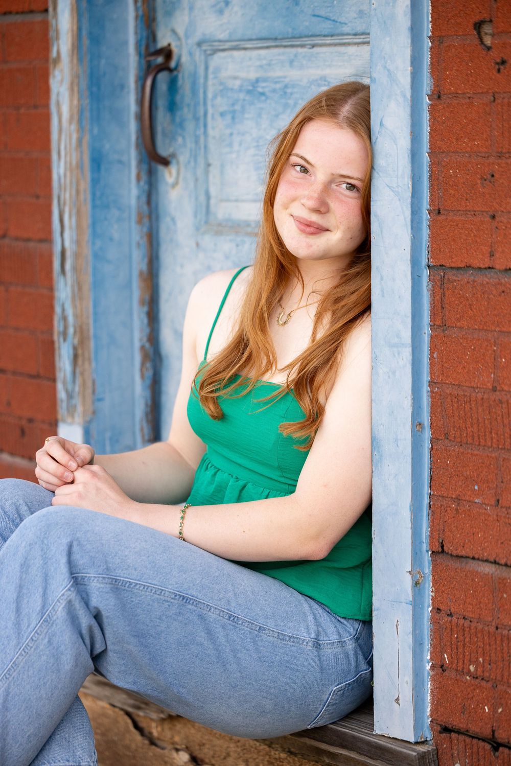 A young woman with long red hair sits in a doorway and smiles at the camera.