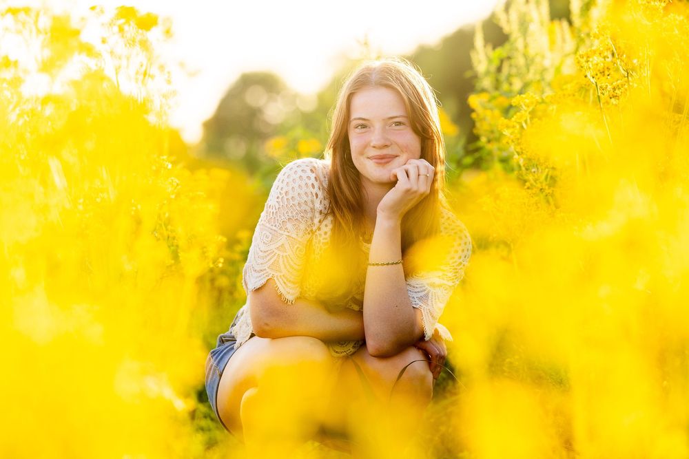 A young woman in a green dress sits among yellow flowers and smiles at the camera.