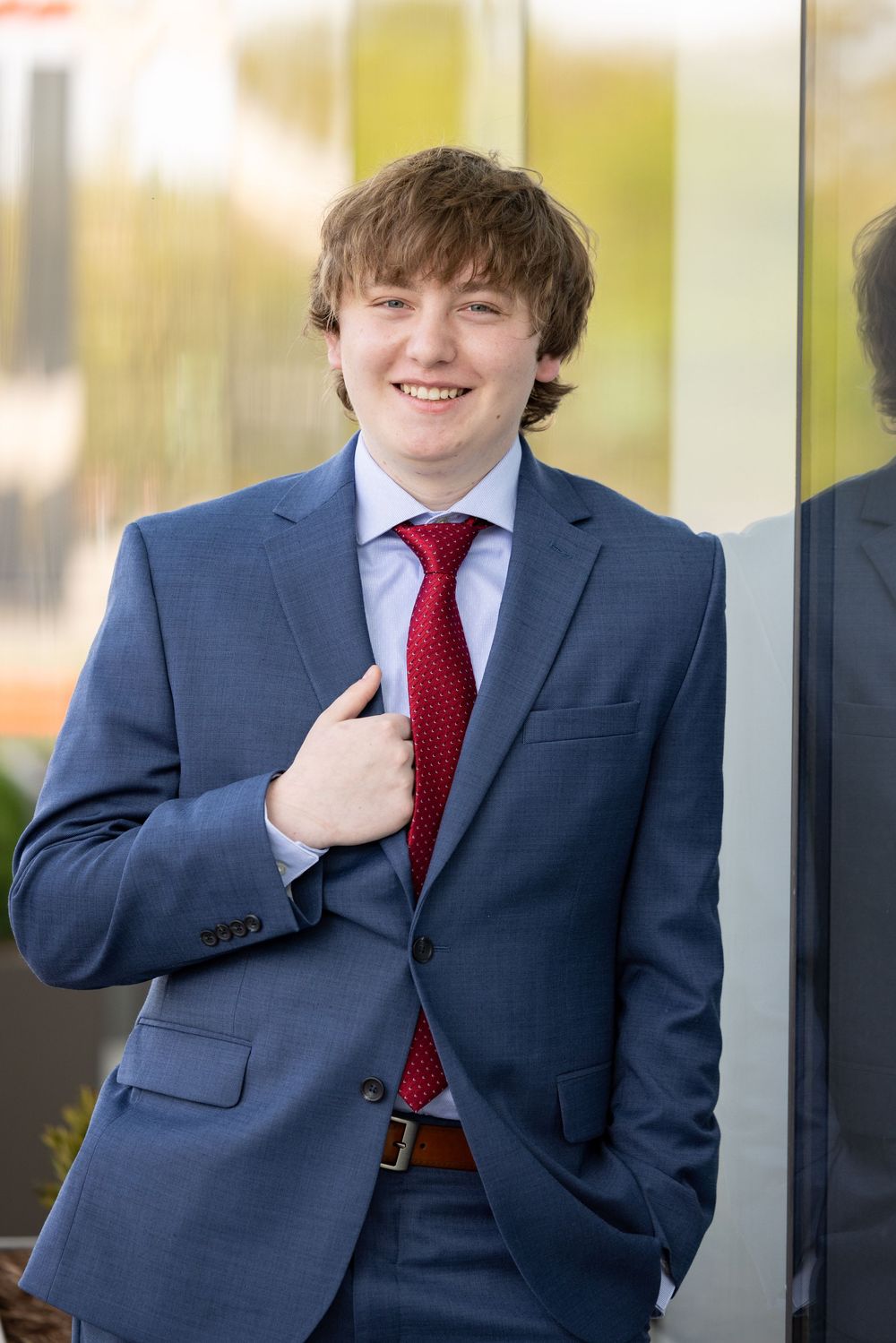 A young man in a suit and tie smiles while standing in front of a blurred background.