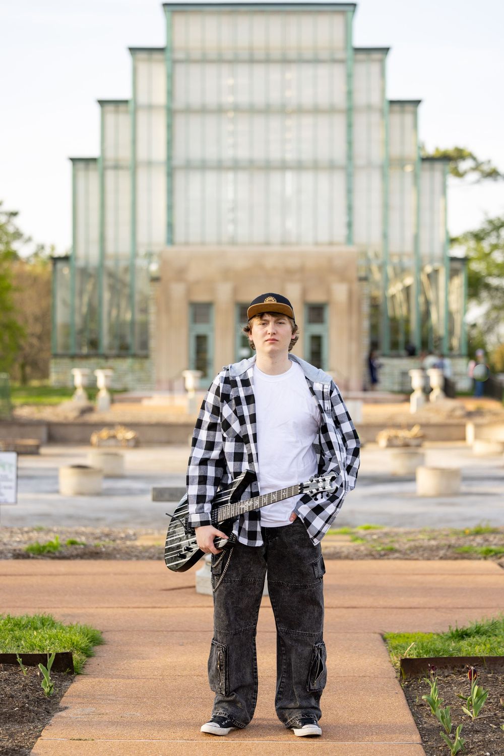 A young man wearing a plaid shirt and baseball cap stands in front of a large glass building.