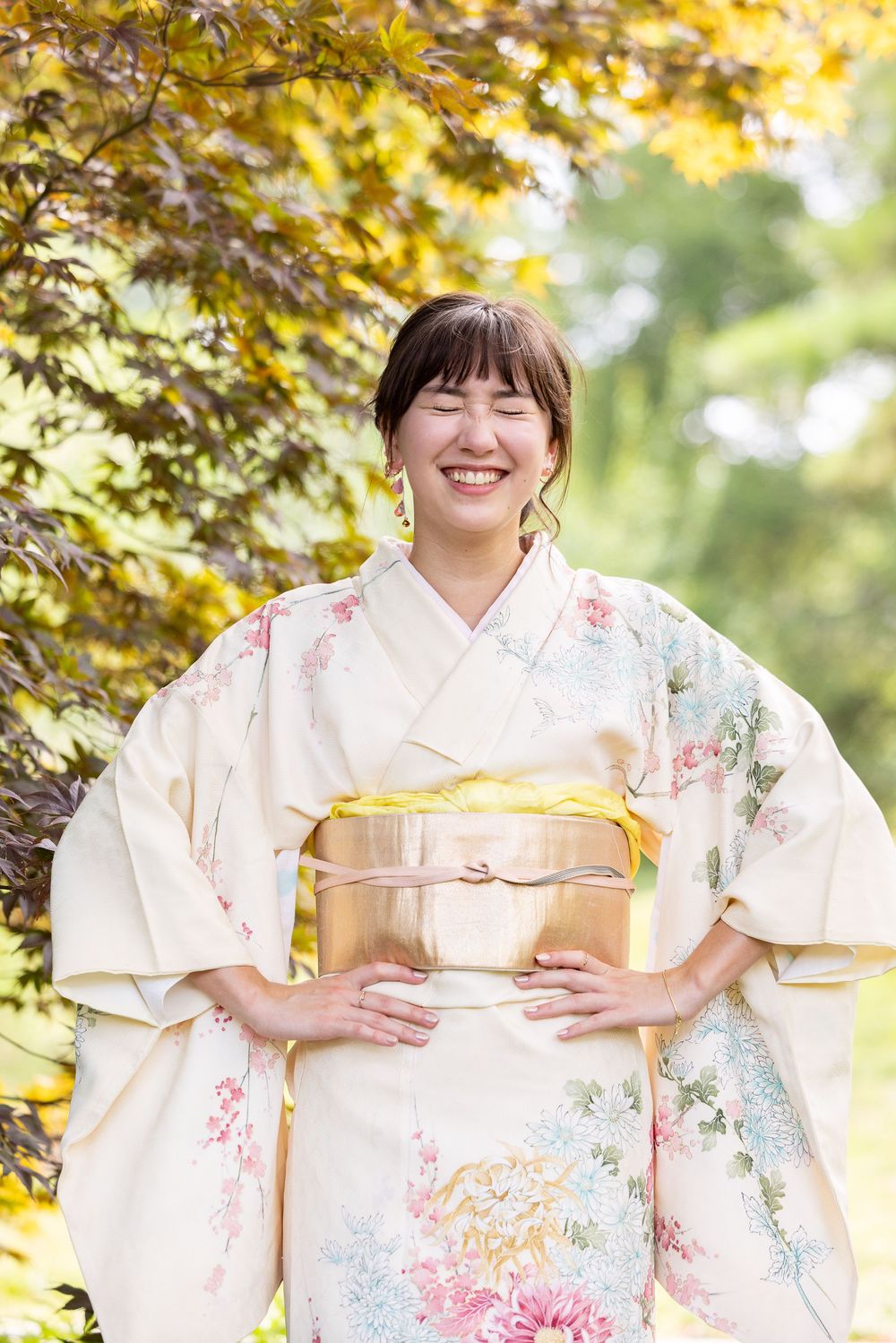 A woman in a colorful kimono holds a traditional Japanese box and smiles in a garden setting.