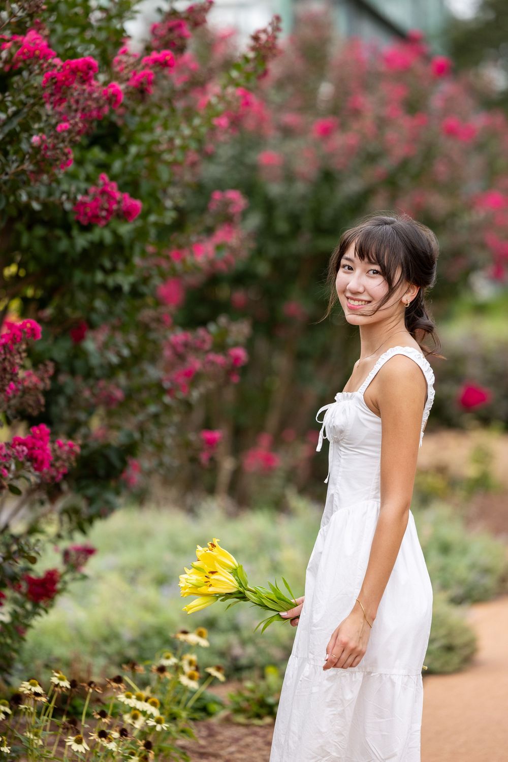 A young woman in a white dress stands among pink flowering bushes and smiles while holding a bouquet of yellow flowers.