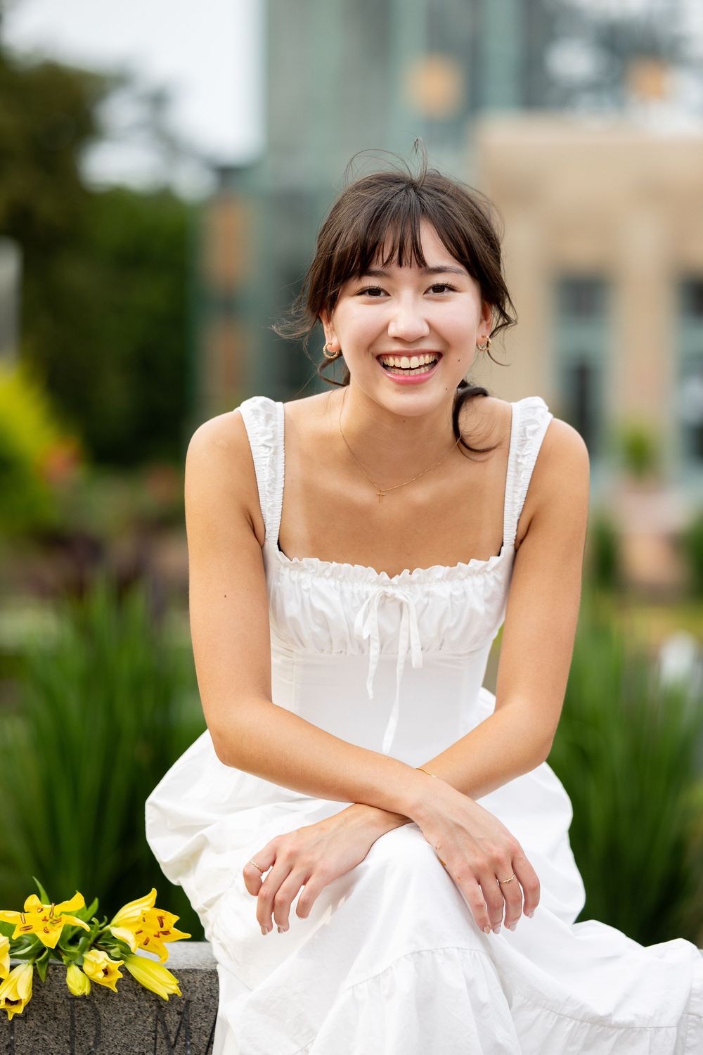 A smiling young woman in a white dress sits on a stone wall in a garden setting.