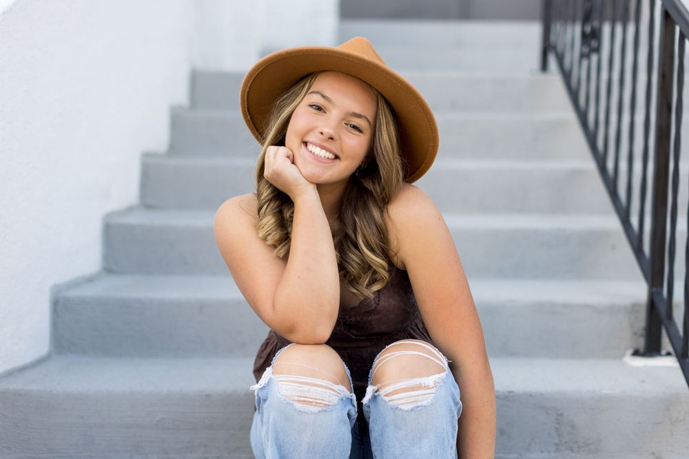 A young woman with long brown hair sits on stairs, smiling and resting her chin on her hands.