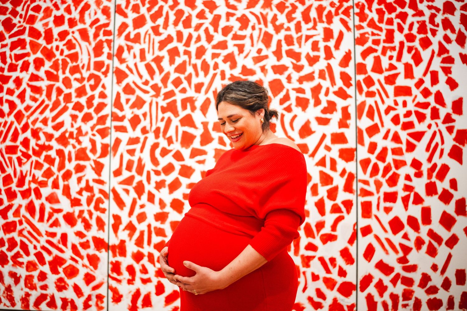 A person in a bright red sweater stands against an abstract red and white patterned wall during a maternity photoshoot.